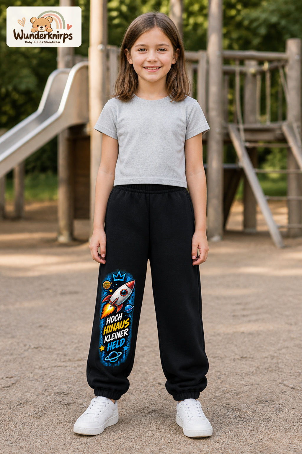 Mädchen trägt schwarze Jogginghose mit Kleiner Held Weltraumrakete-Print, von vorn auf dem Spielplatz.