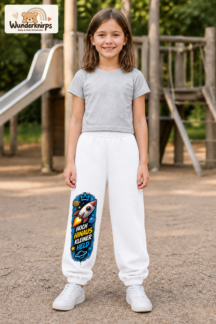 Mädchen trägt weiße Jogginghose mit Kleiner Held Weltraumrakete-Print, von vorn auf dem Spielplatz.