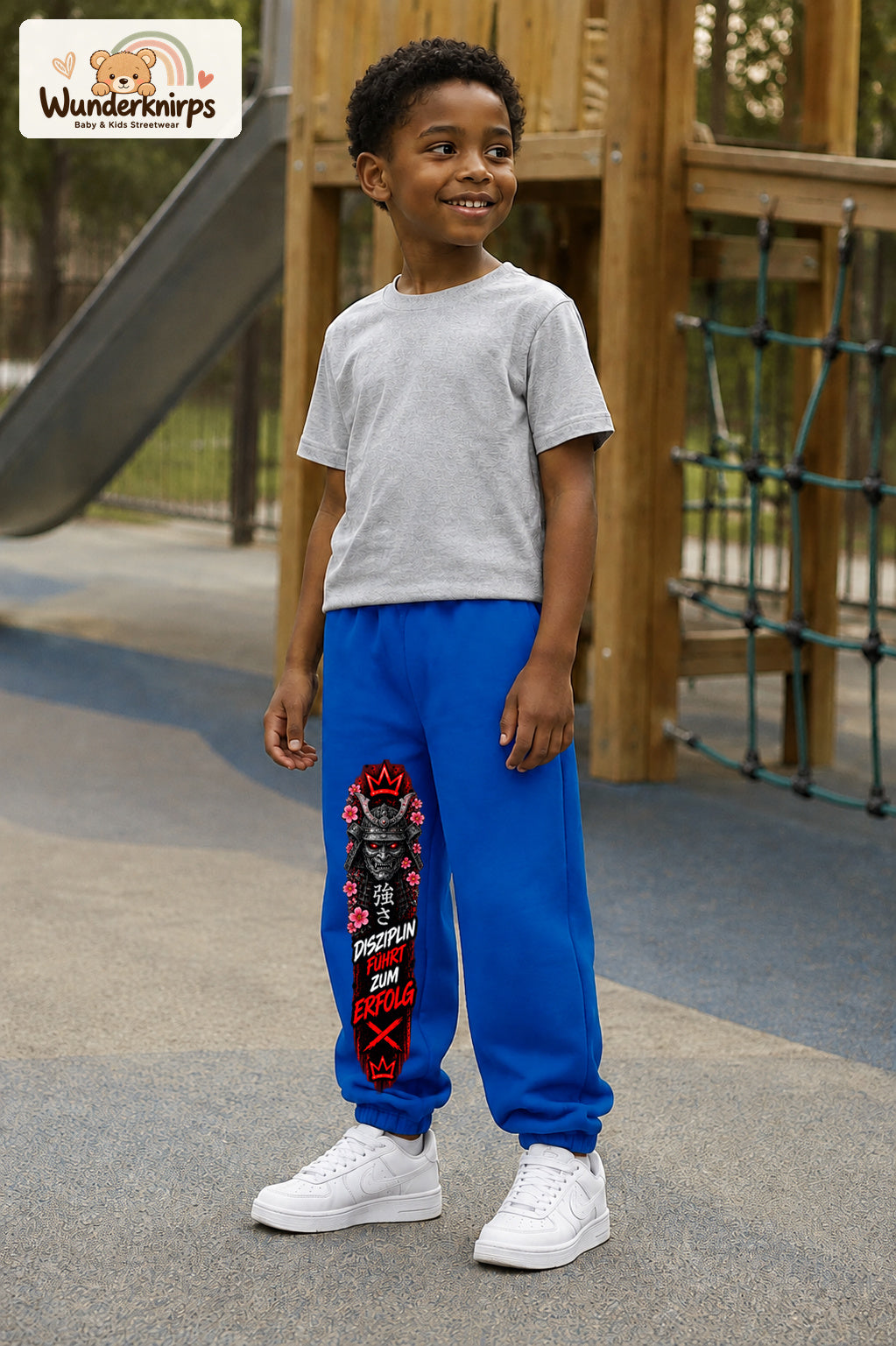 Junge trägt blaue Jogginghose mit Disziplin Samurai-Print, leicht seitlich auf dem Spielplatz.