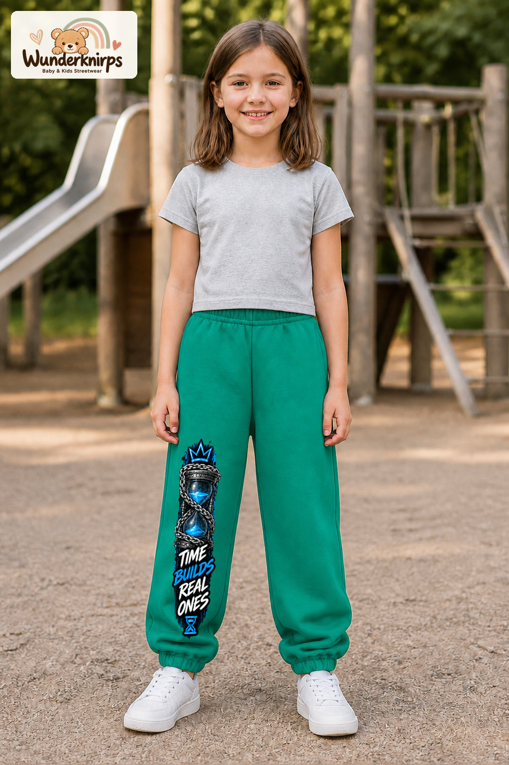 Mädchen trägt grüne Jogginghose mit Time Builds Sanduhr-Print, von vorn auf dem Spielplatz.