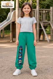 Mädchen trägt grüne Jogginghose mit Time Builds Sanduhr-Print, von vorn auf dem Spielplatz.