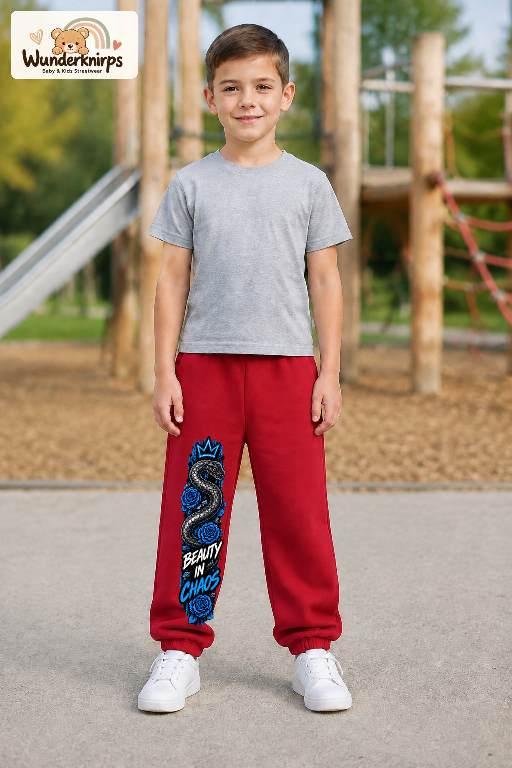 Junge trägt rote Jogginghose mit Beauty in Chaos Rosen-Print, von vorn auf dem Spielplatz.