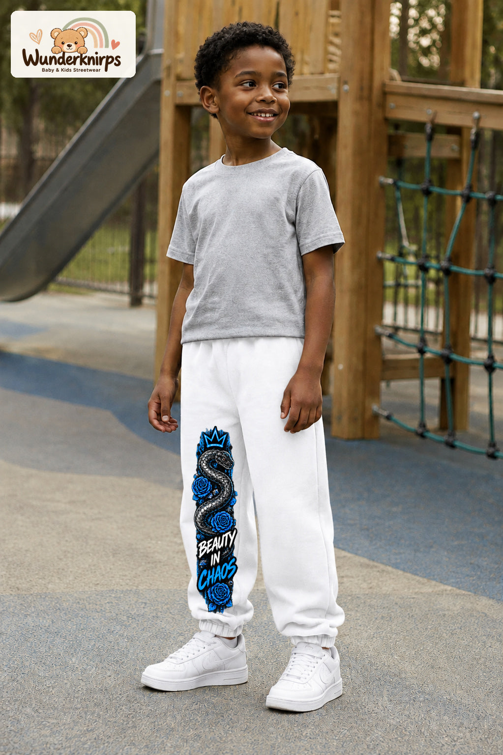 Junge trägt weiße Jogginghose mit Beauty in Chaos Rosen-Print, leicht seitlich auf dem Spielplatz.