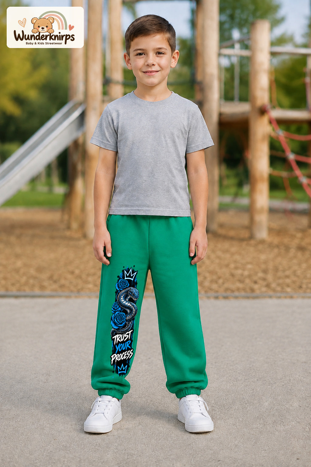 Junge trägt grüne Jogginghose mit Trust Your Process Schlange-Print, von vorn auf dem Spielplatz.