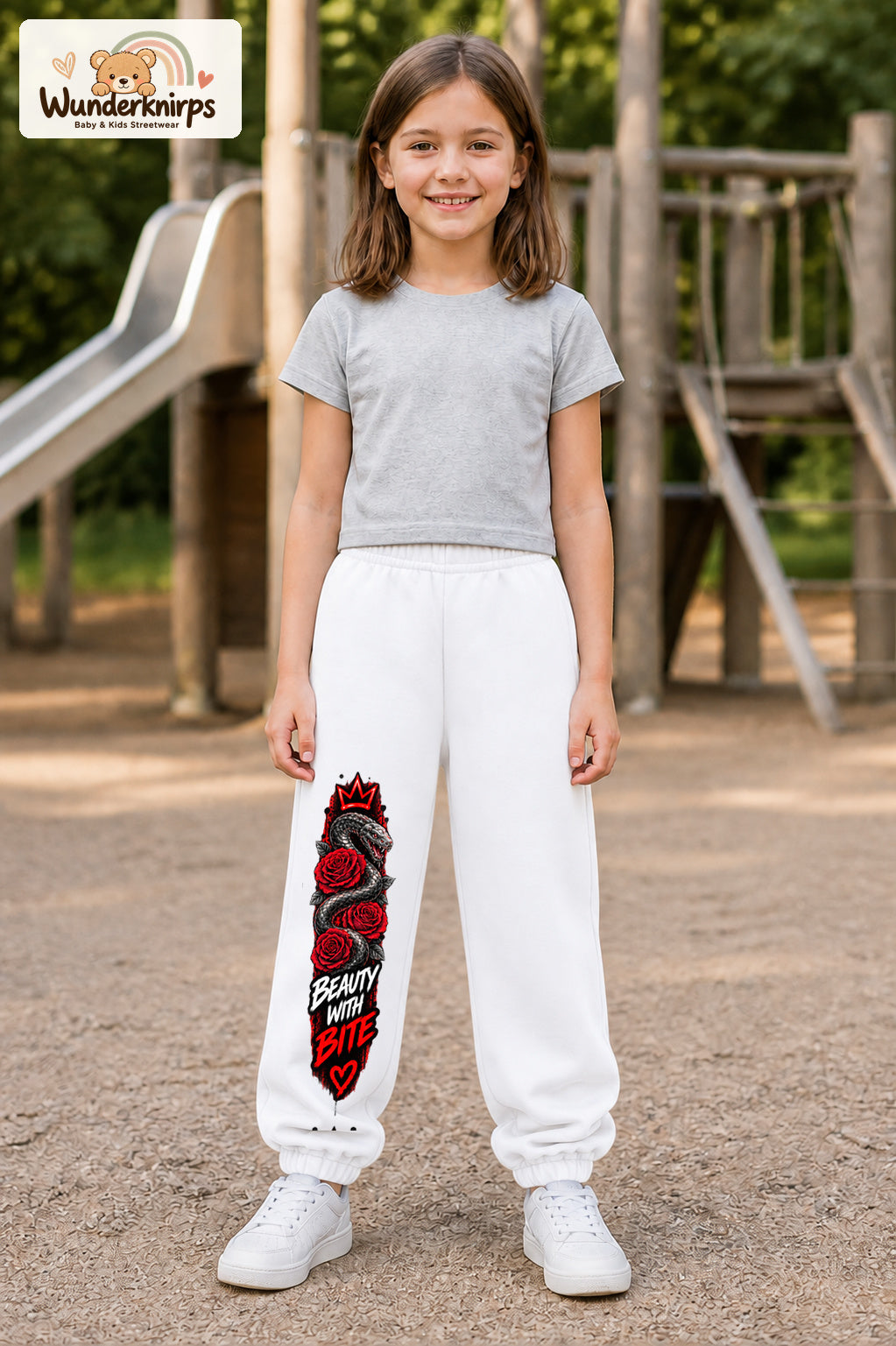 Mädchen trägt weiße Jogginghose mit Beauty with Bite-Print, von vorn auf dem Spielplatz.