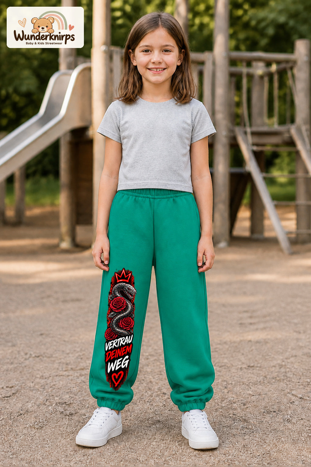 Mädchen trägt grüne Jogginghose mit Vertrau deinem Weg-Print, von vorn auf dem Spielplatz.
