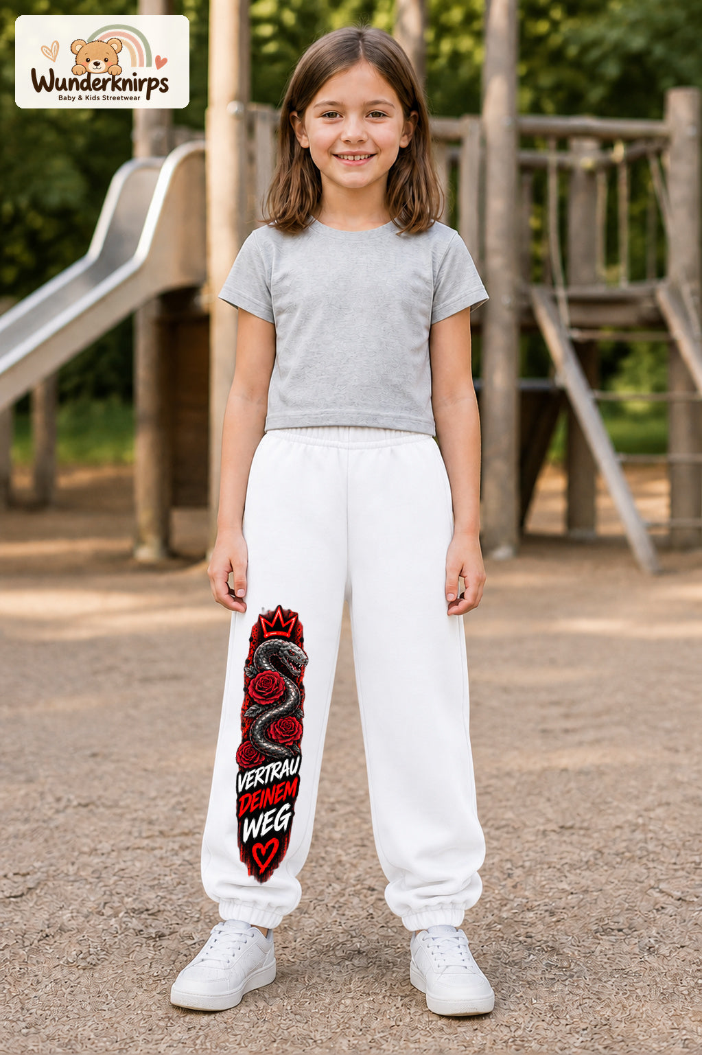 Mädchen trägt weiße Jogginghose mit Vertrau deinem Weg-Print, von vorn auf dem Spielplatz.