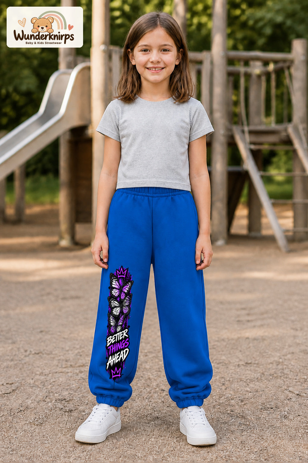 Mädchen trägt blaue Jogginghose mit Better Things Falter-Print, von vorn auf dem Spielplatz.