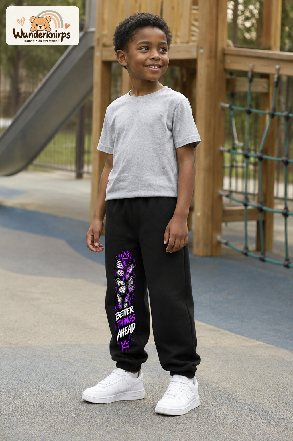 Junge trägt schwarze Jogginghose mit Better Things Falter-Print, leicht seitlich auf dem Spielplatz.