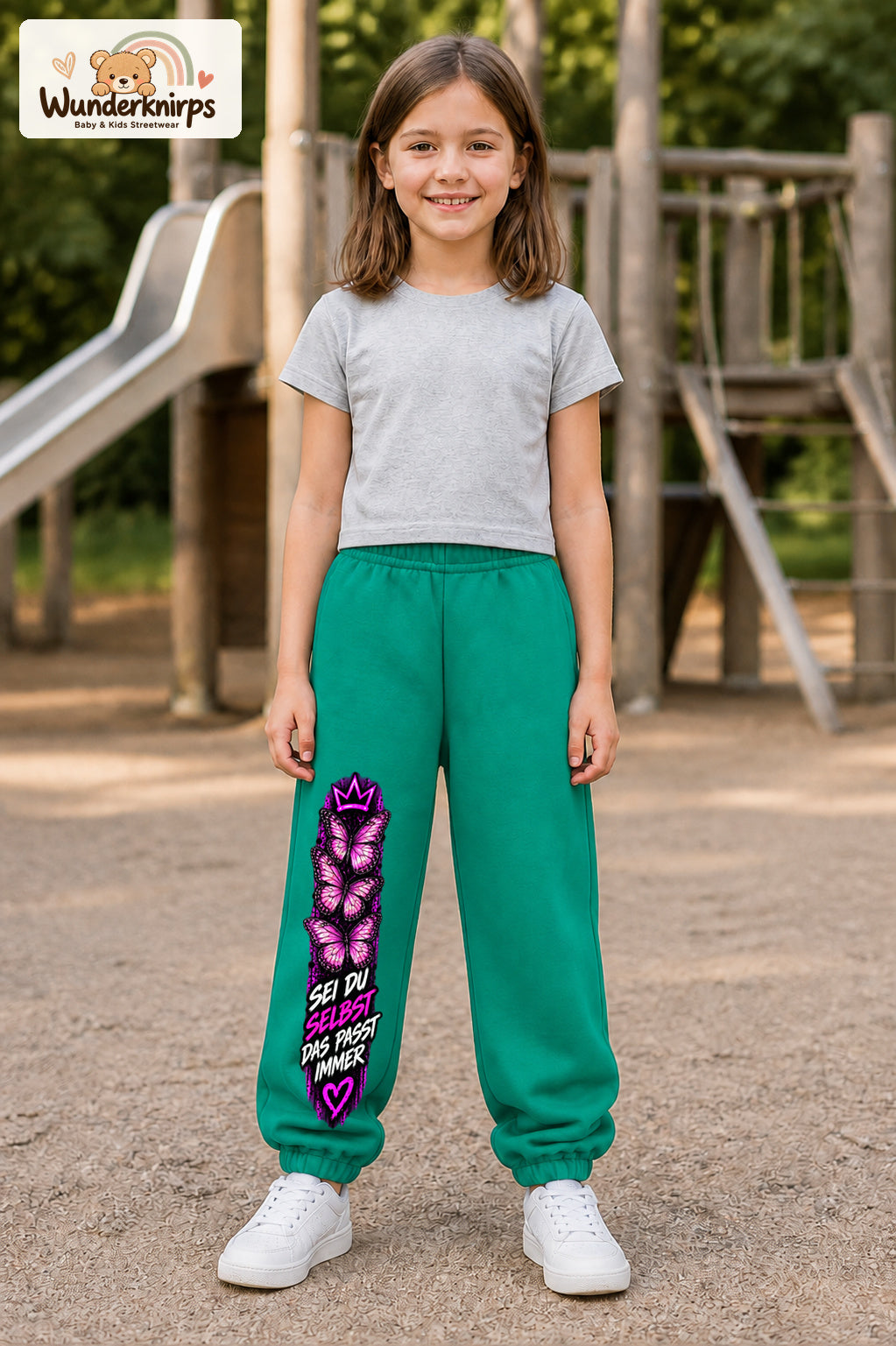Mädchen trägt grüne Jogginghose mit Sei du selbst Falter-Print, von vorn auf dem Spielplatz.