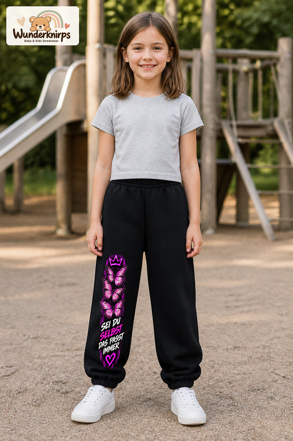 Mädchen trägt schwarze Jogginghose mit Sei du selbst Falter-Print, von vorn auf dem Spielplatz.
