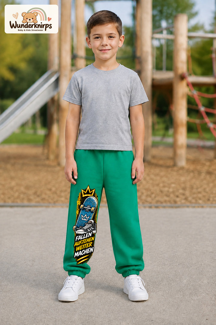 Junge trägt grüne Jogginghose mit Aufstehen Skateboard-Print, von vorn auf dem Spielplatz.