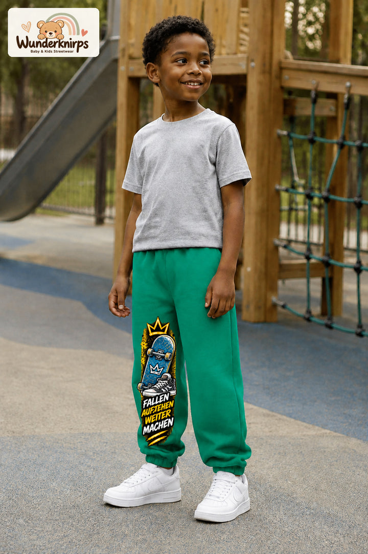 Junge trägt grüne Jogginghose mit Aufstehen Skateboard-Print, leicht seitlich auf dem Spielplatz.