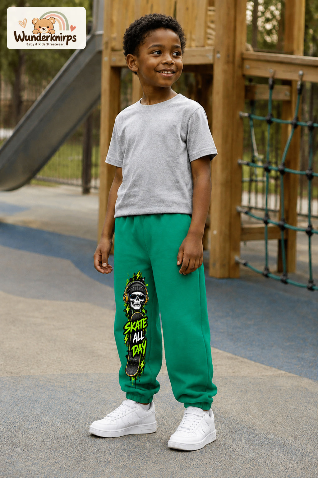 Junge trägt grüne Jogginghose mit Skate All Day Skull-Print, leicht seitlich auf dem Spielplatz.