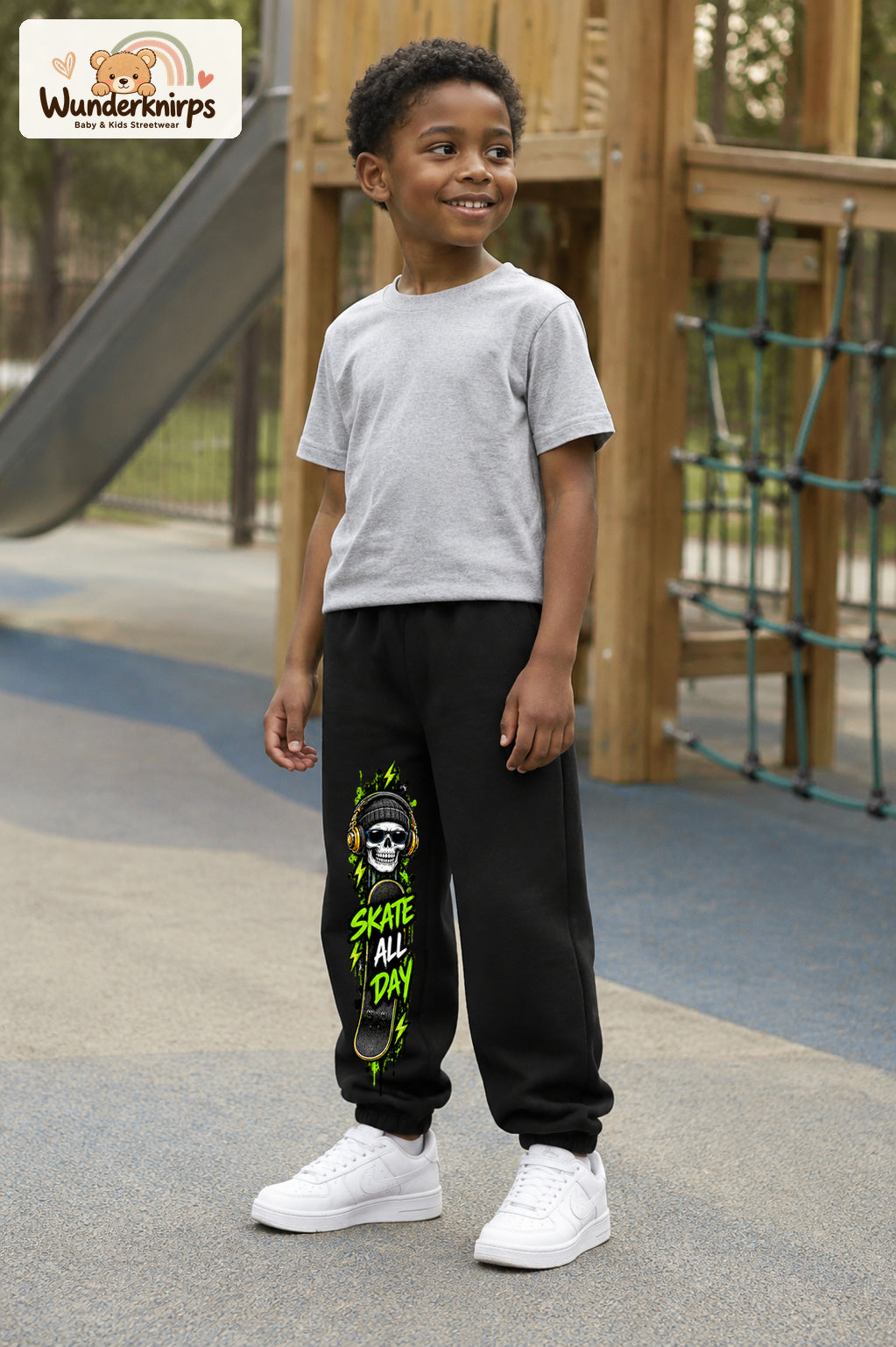 Junge trägt schwarze Jogginghose mit Skate All Day Skull-Print, leicht seitlich auf dem Spielplatz.
