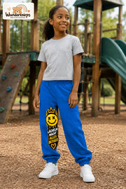 Mädchen trägt blaue Jogginghose mit Lächeln macht besser-Print, leicht seitlich auf dem Spielplatz.