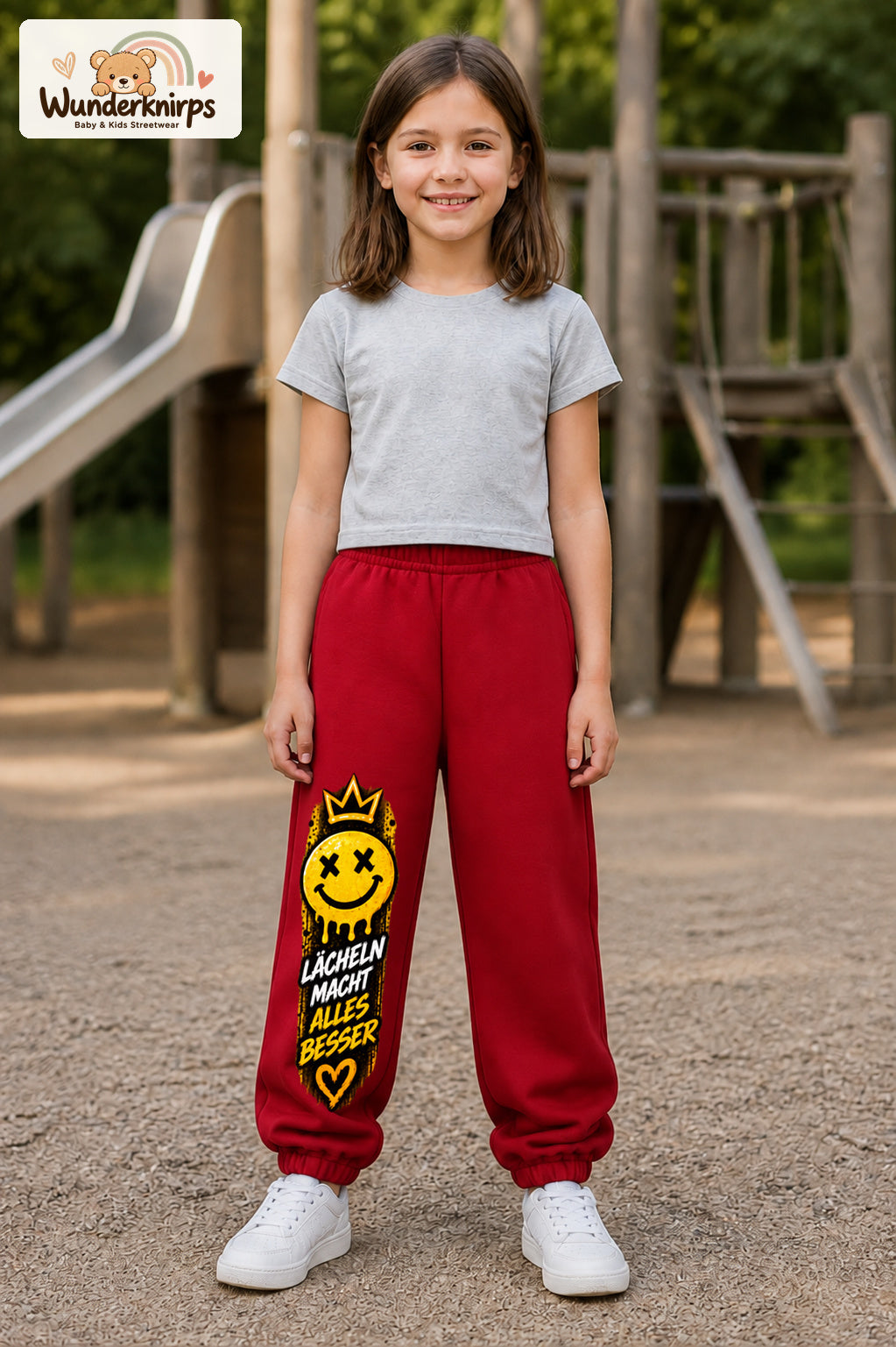 Mädchen trägt rote Jogginghose mit Lächeln macht besser-Print, von vorn auf dem Spielplatz.