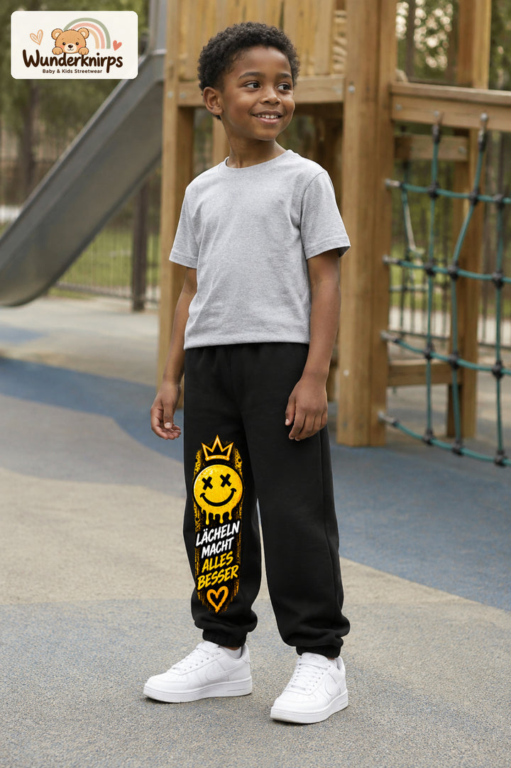 Junge trägt schwarze Jogginghose mit Lächeln macht besser-Print, leicht seitlich auf dem Spielplatz.