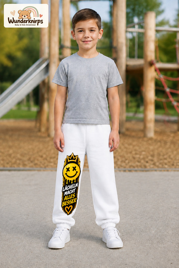 Junge trägt weiße Jogginghose mit Lächeln macht besser-Print, von vorn auf dem Spielplatz.