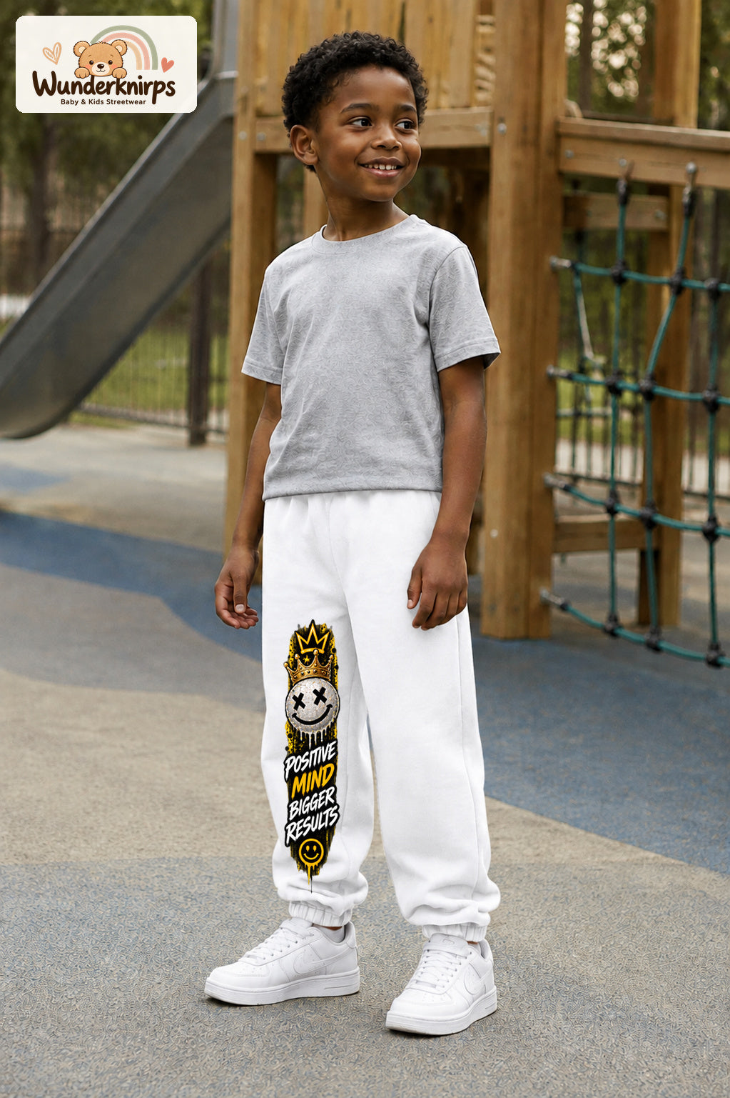 Junge trägt weiße Jogginghose mit Positive Mind Smiley-Print, leicht seitlich auf dem Spielplatz.