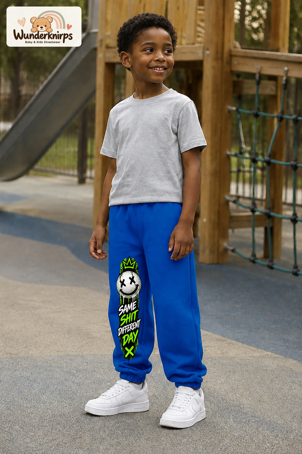 Junge trägt blaue Jogginghose mit Same Shit Smiley-Print, leicht seitlich auf dem Spielplatz.