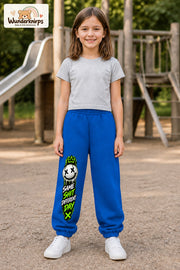 Mädchen trägt blaue Jogginghose mit Same Shit Smiley-Print, von vorn auf dem Spielplatz.