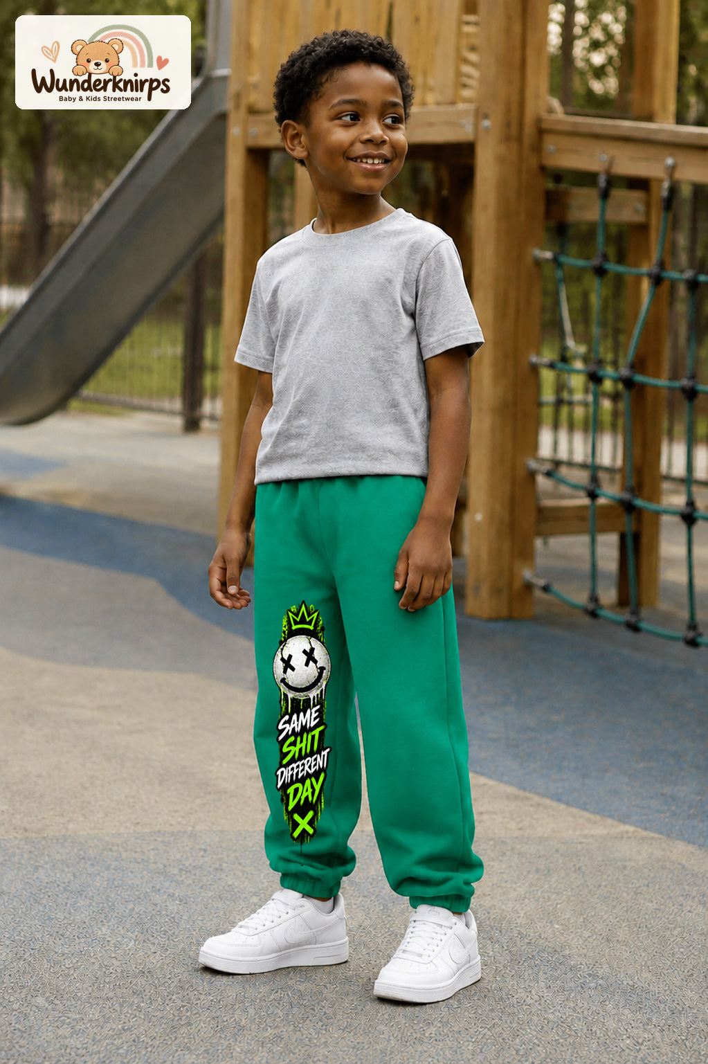Junge trägt grüne Jogginghose mit Same Shit Smiley-Print, leicht seitlich auf dem Spielplatz.