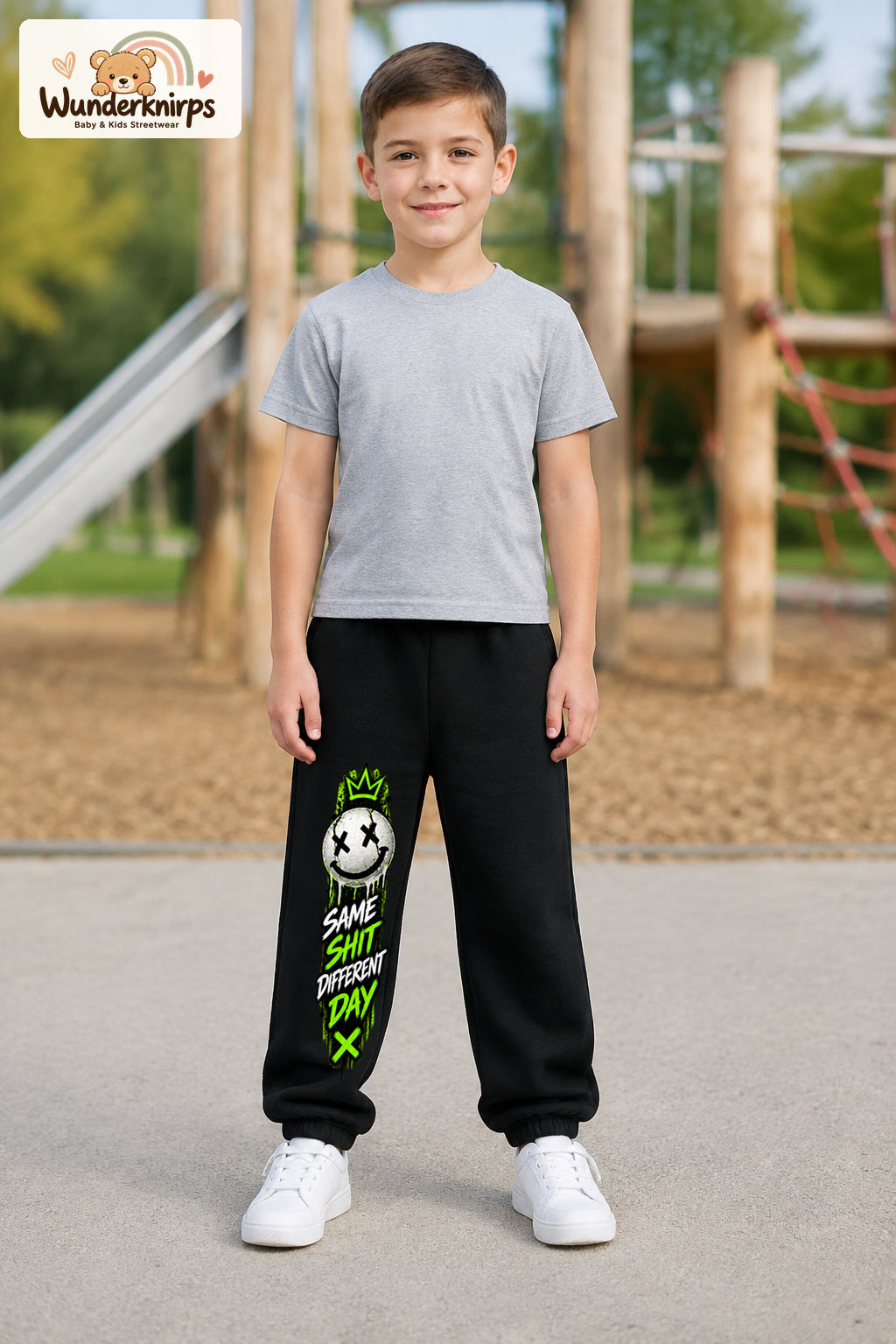 Junge trägt schwarze Jogginghose mit Same Shit Smiley-Print, von vorn auf dem Spielplatz.