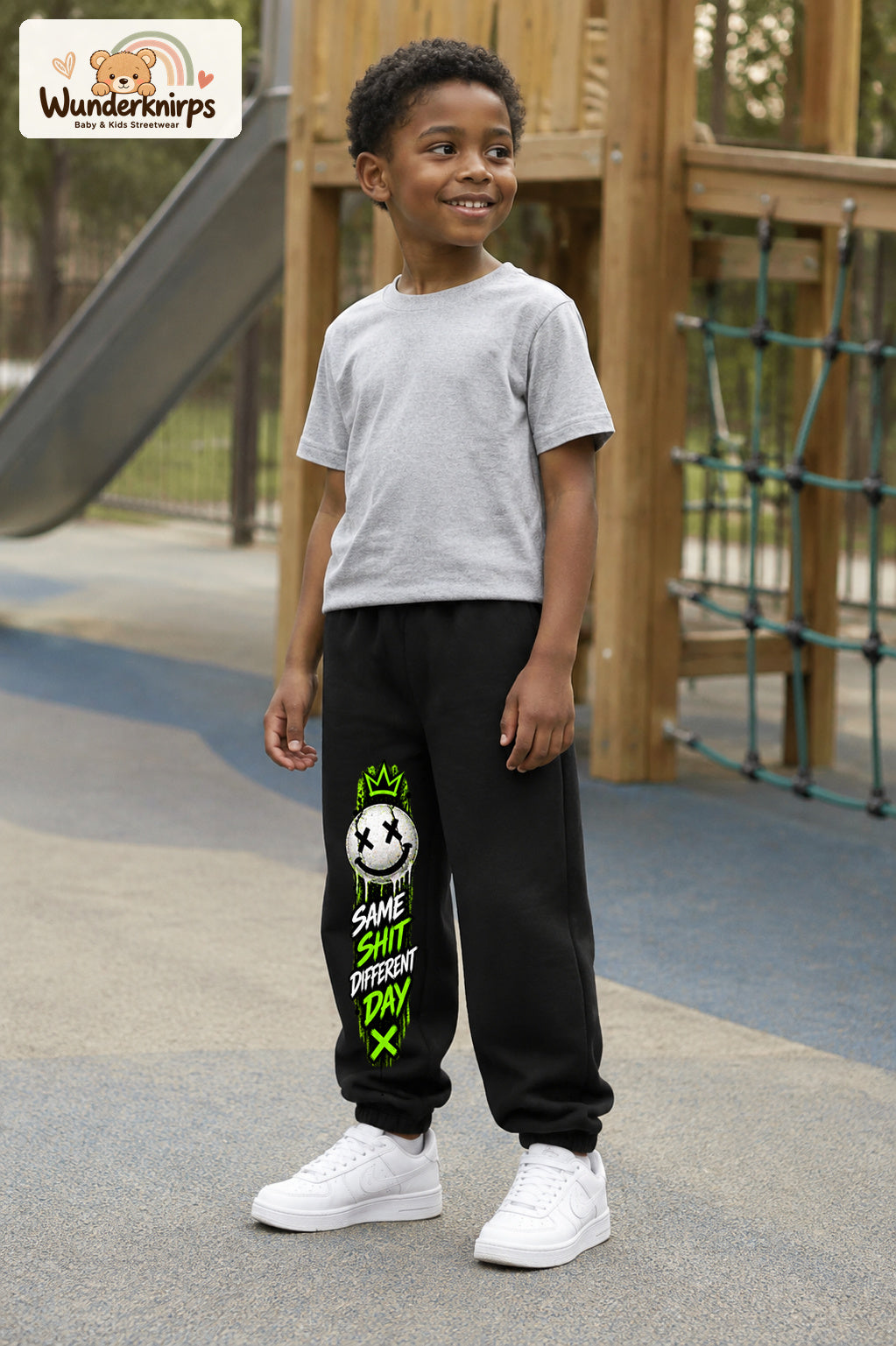 Junge trägt schwarze Jogginghose mit Same Shit Smiley-Print, leicht seitlich auf dem Spielplatz.
