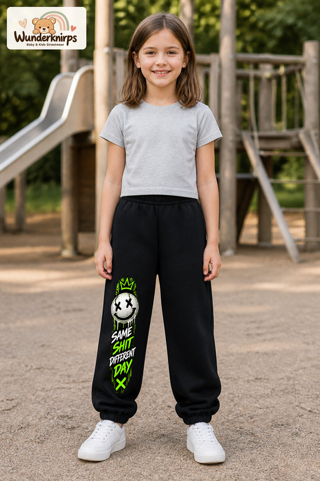Mädchen trägt schwarze Jogginghose mit Same Shit Smiley-Print, von vorn auf dem Spielplatz.
