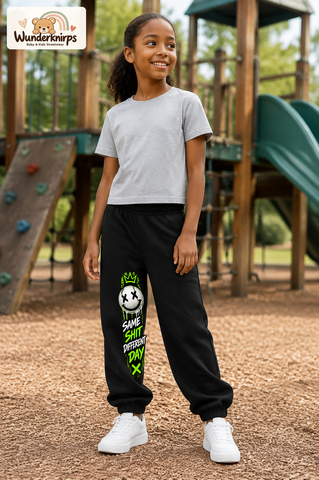 Mädchen trägt schwarze Jogginghose mit Same Shit Smiley-Print, leicht seitlich auf dem Spielplatz.