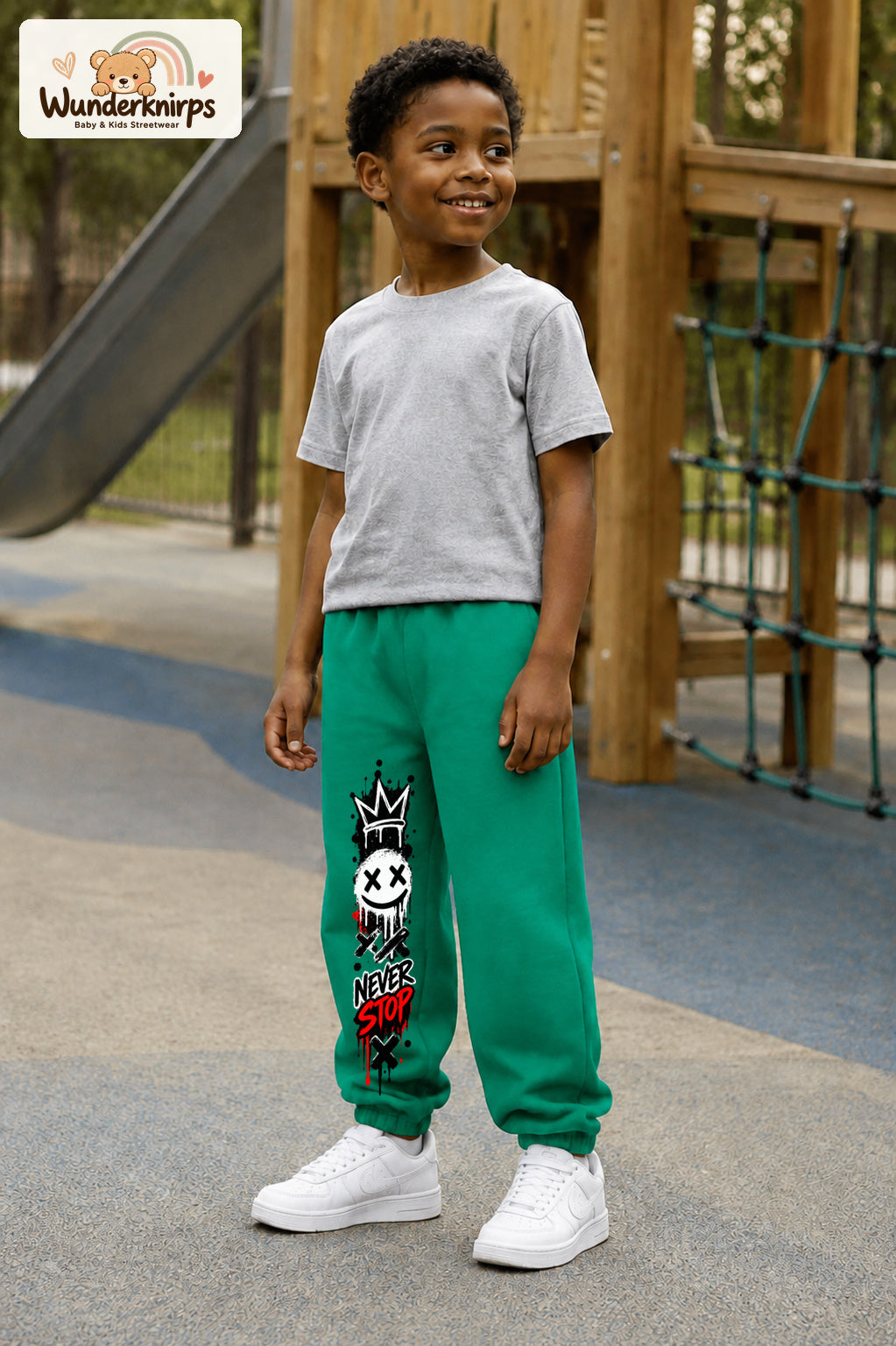 Junge trägt grüne Jogginghose mit Never Stop Smiley-Print, leicht seitlich auf dem Spielplatz.