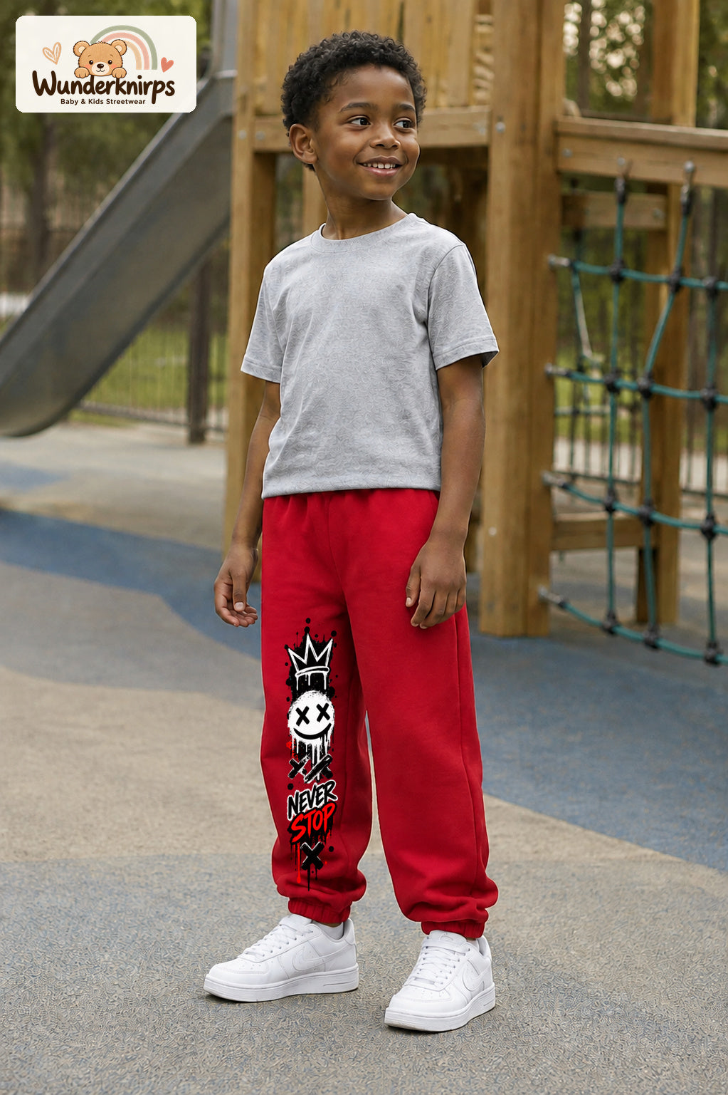 Junge trägt rote Jogginghose mit Never Stop Smiley-Print, leicht seitlich auf dem Spielplatz.