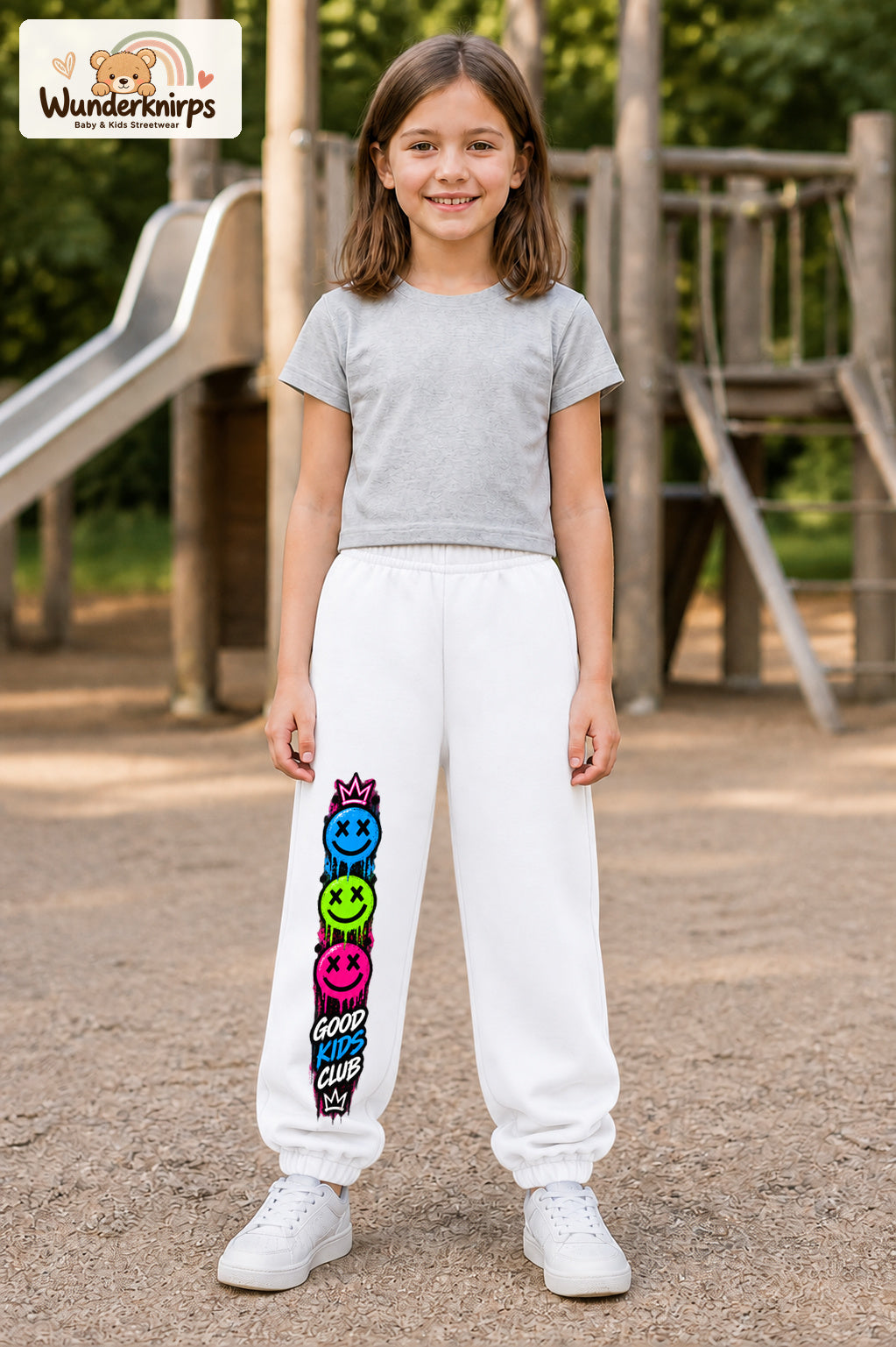 Mädchen trägt weiße Jogginghose mit Good Kids Smileys-Print, von vorn auf dem Spielplatz.
