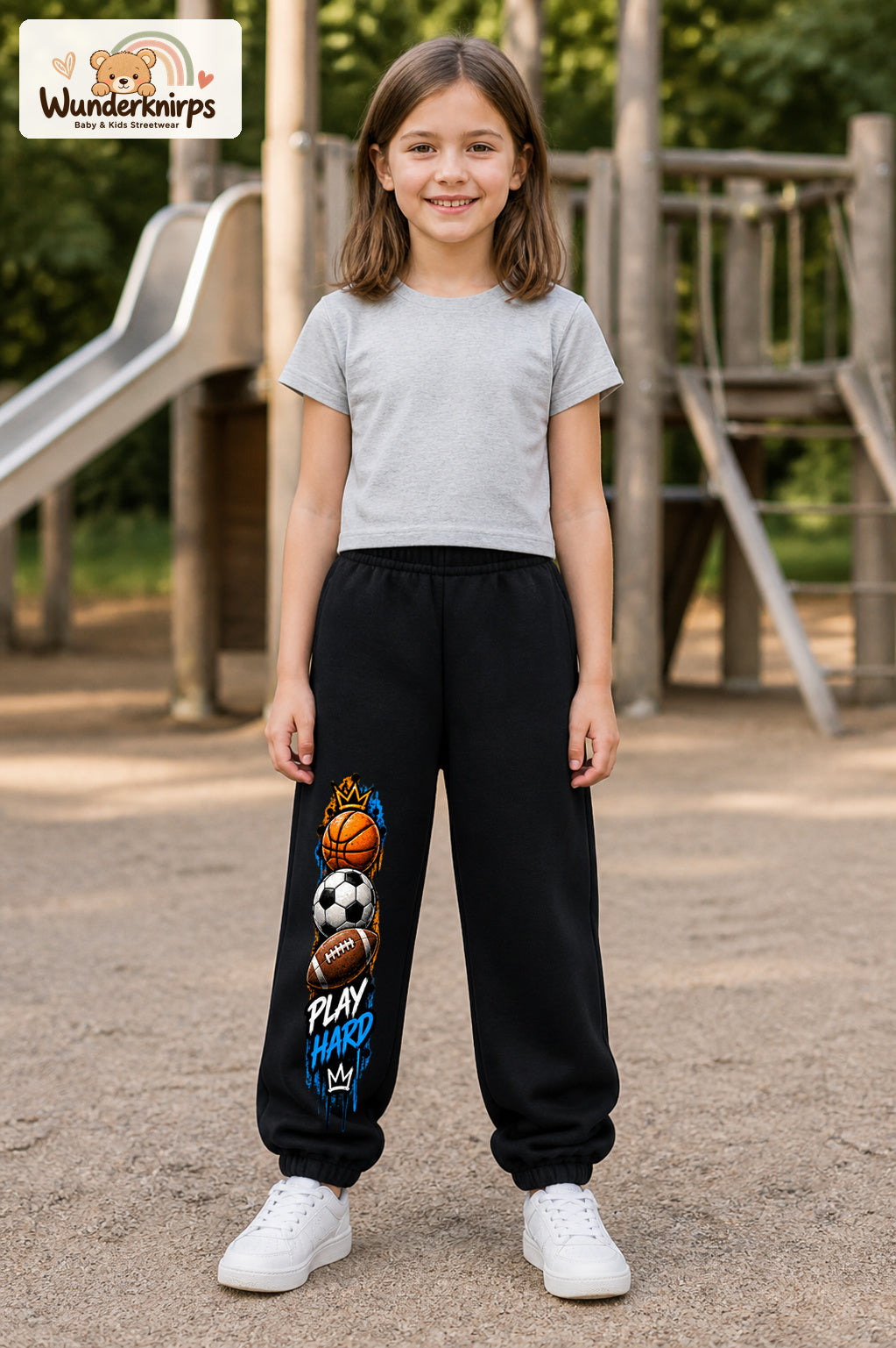 Mädchen trägt schwarze Jogginghose mit Play Hard Sportmix-Print, von vorn auf dem Spielplatz.