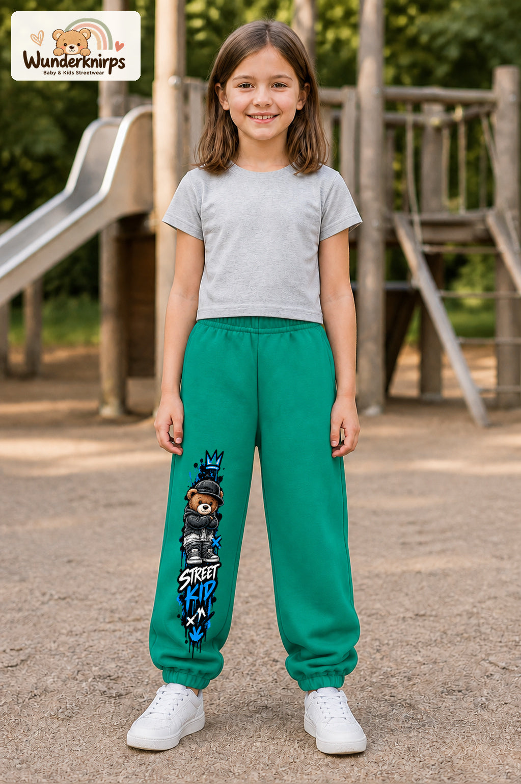 Mädchen trägt grüne Jogginghose mit Street Kid Teddy-Print, von vorn auf dem Spielplatz.