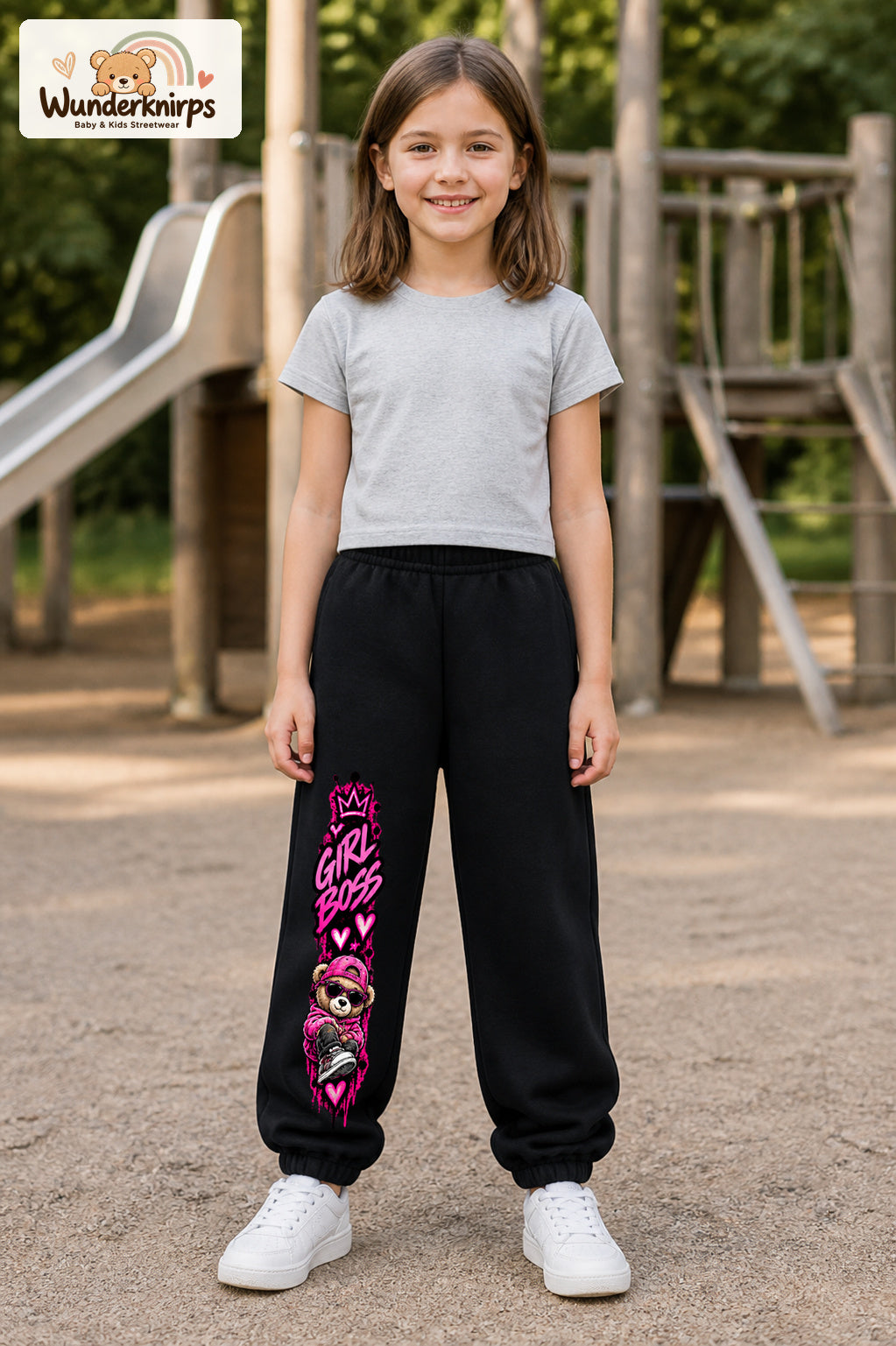 Mädchen trägt schwarze Jogginghose mit Girl Boss Bär-Print, von vorn auf dem Spielplatz.