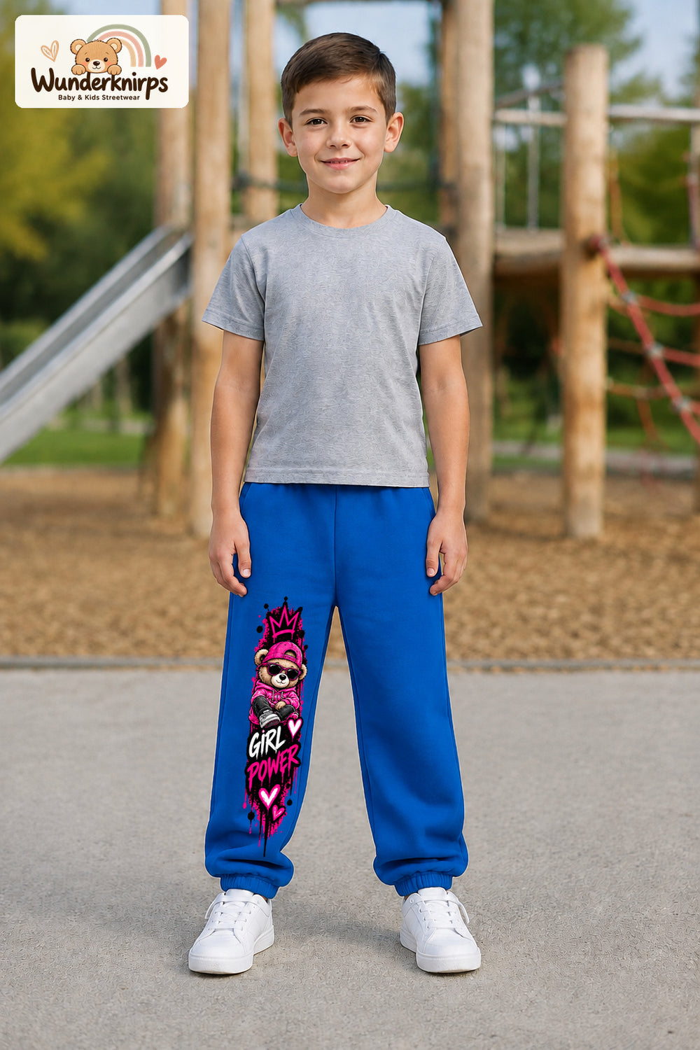 Junge trägt blaue Jogginghose mit Girl Power Teddy-Print, von vorn auf dem Spielplatz.