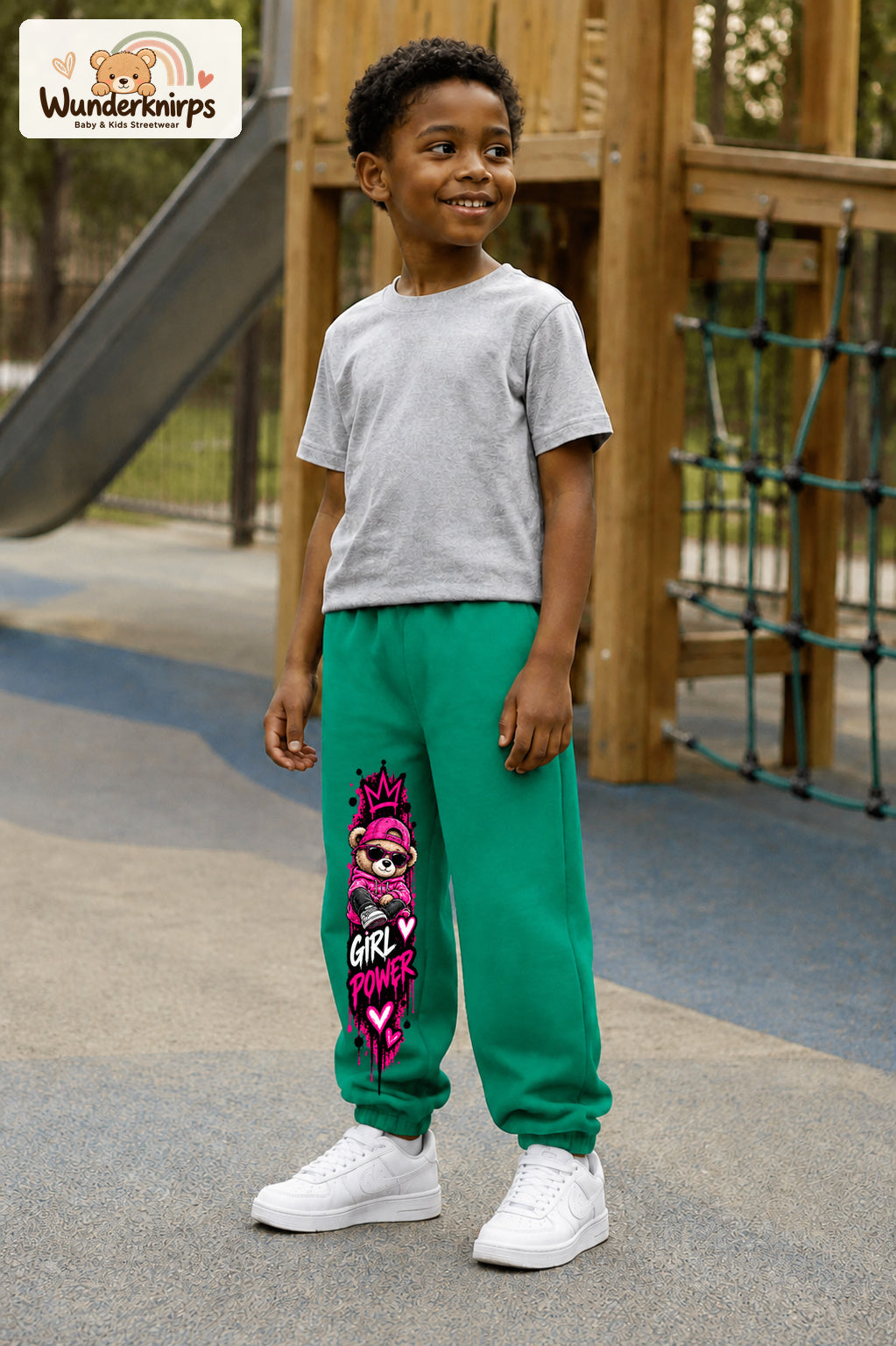 Junge trägt grüne Jogginghose mit Girl Power Teddy-Print, leicht seitlich auf dem Spielplatz.
