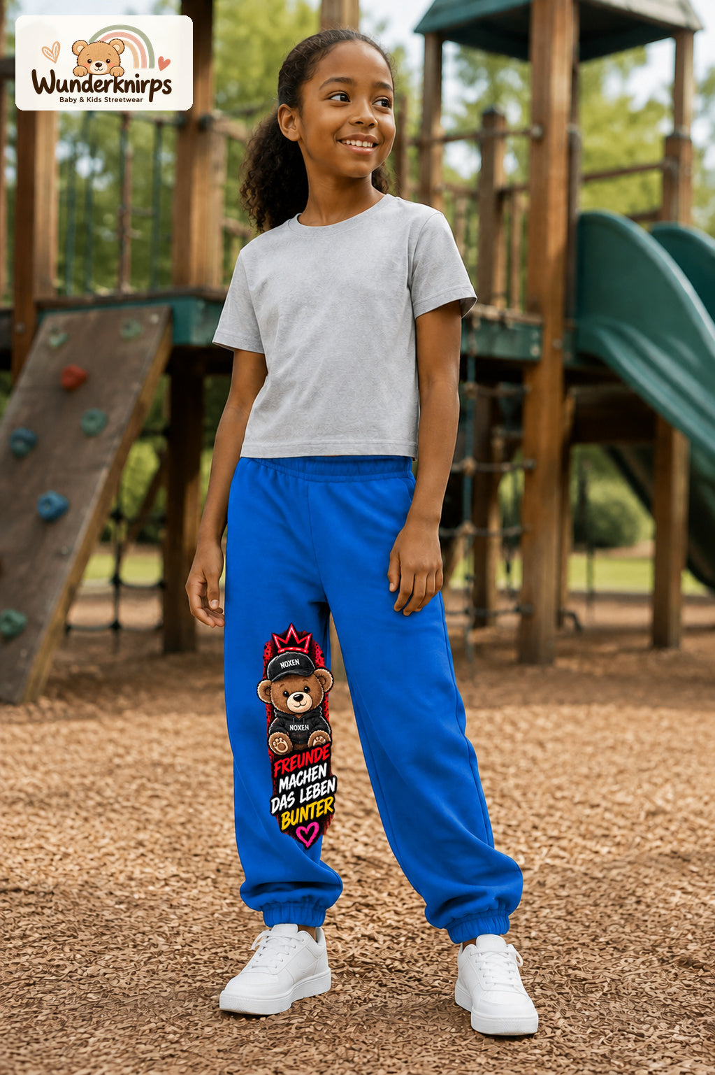 Mädchen trägt blaue Jogginghose mit Freunde machen bunt Teddy-Print, leicht seitlich auf dem Spielplatz.