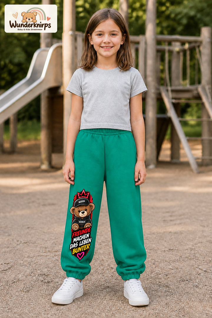 Mädchen trägt grüne Jogginghose mit Freunde machen bunt Teddy-Print, von vorn auf dem Spielplatz.