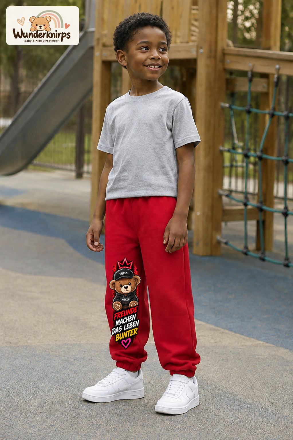 Junge trägt rote Jogginghose mit Freunde machen bunt Teddy-Print, leicht seitlich auf dem Spielplatz.