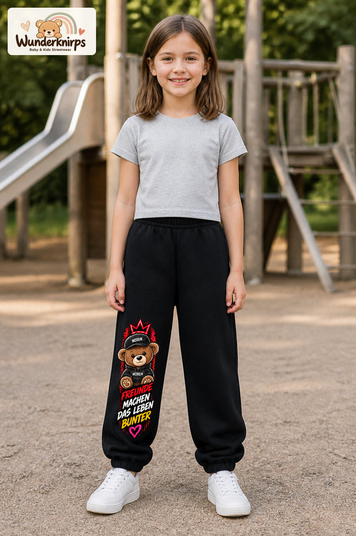Mädchen trägt schwarze Jogginghose mit Freunde machen bunt Teddy-Print, von vorn auf dem Spielplatz.