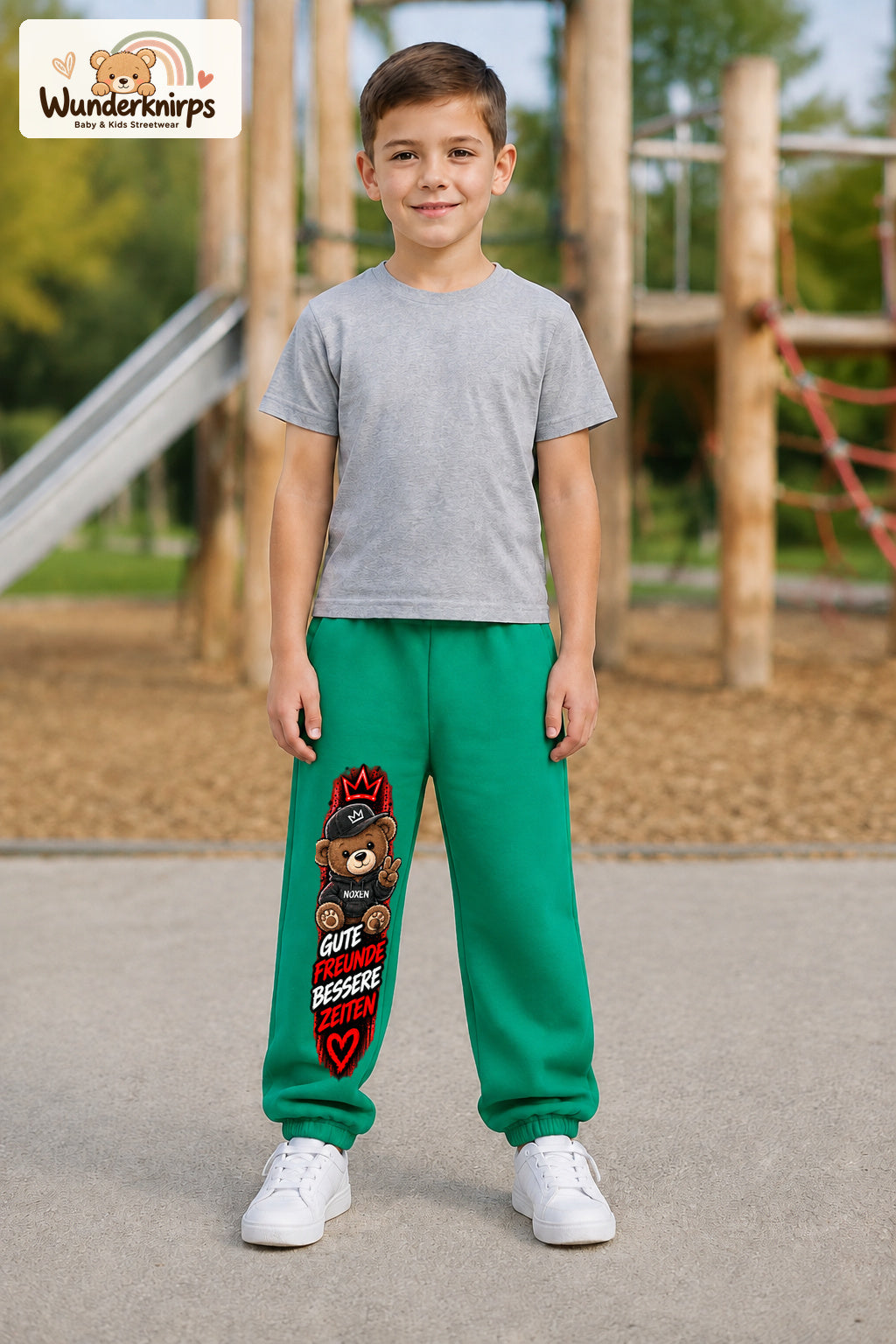 Junge trägt grüne Jogginghose mit Gute Freunde Teddy-Print, von vorn auf dem Spielplatz.