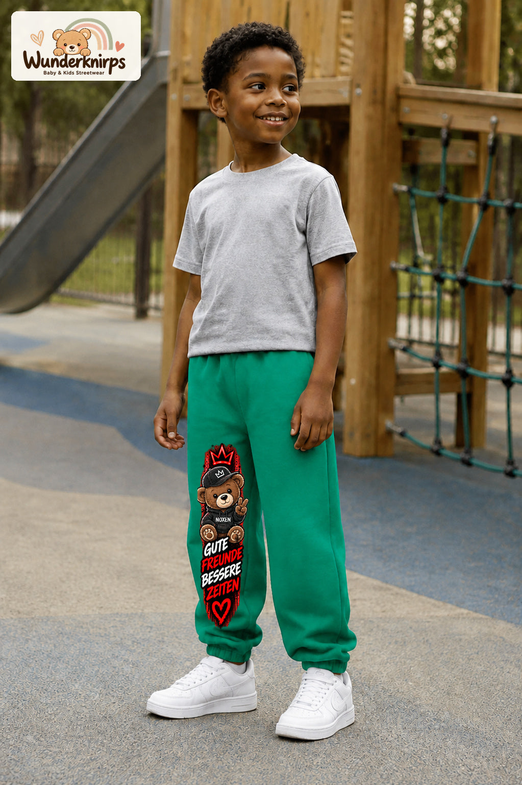 Junge trägt grüne Jogginghose mit Gute Freunde Teddy-Print, leicht seitlich auf dem Spielplatz.