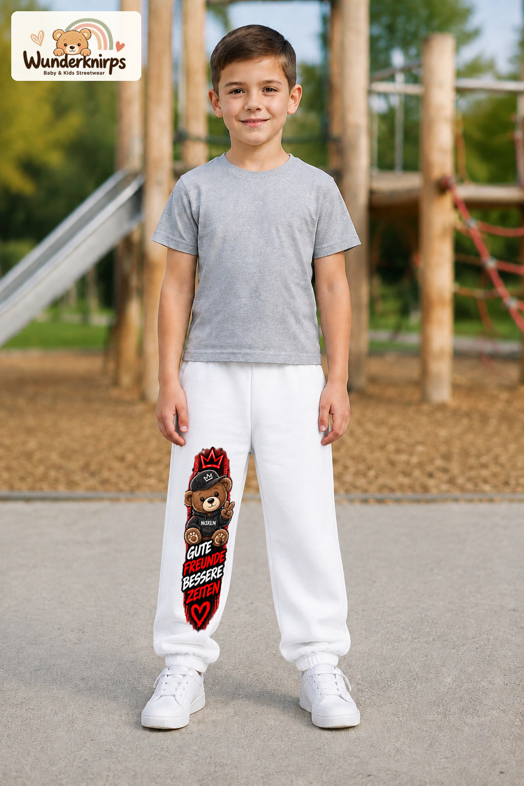 Junge trägt weiße Jogginghose mit Gute Freunde Teddy-Print, von vorn auf dem Spielplatz.