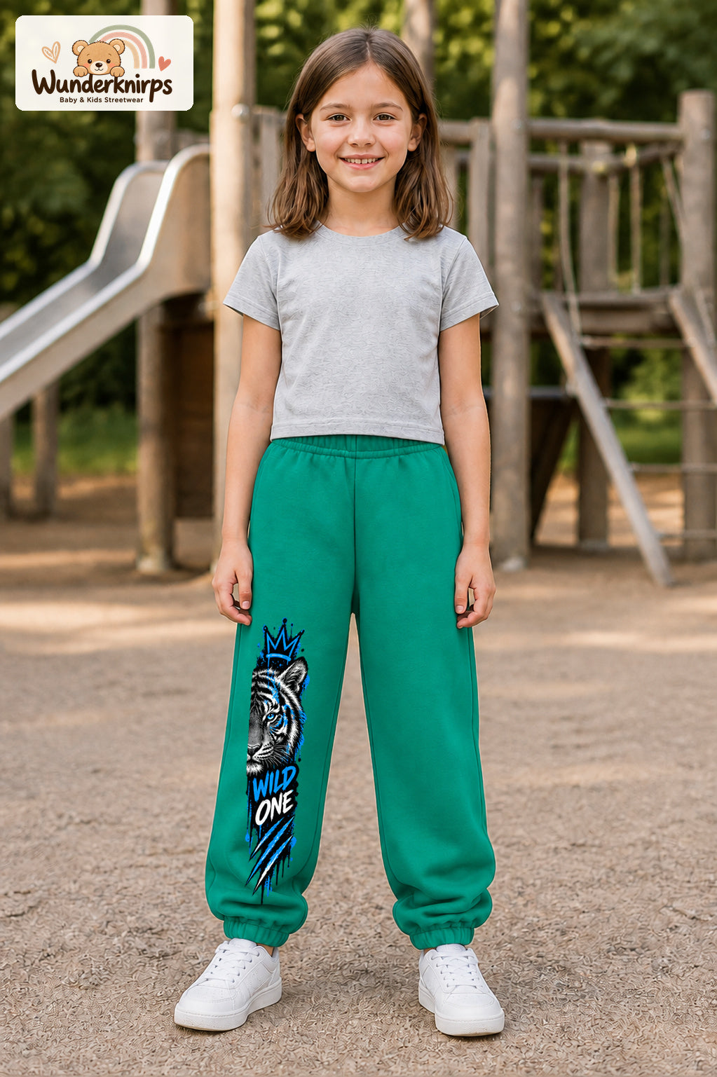 Mädchen trägt grüne Jogginghose mit Wild One Tiger-Print, von vorn auf dem Spielplatz.