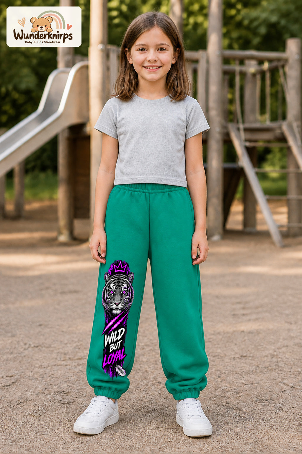 Mädchen trägt grüne Jogginghose mit Wild but Loyal Tiger-Print, von vorn auf dem Spielplatz.