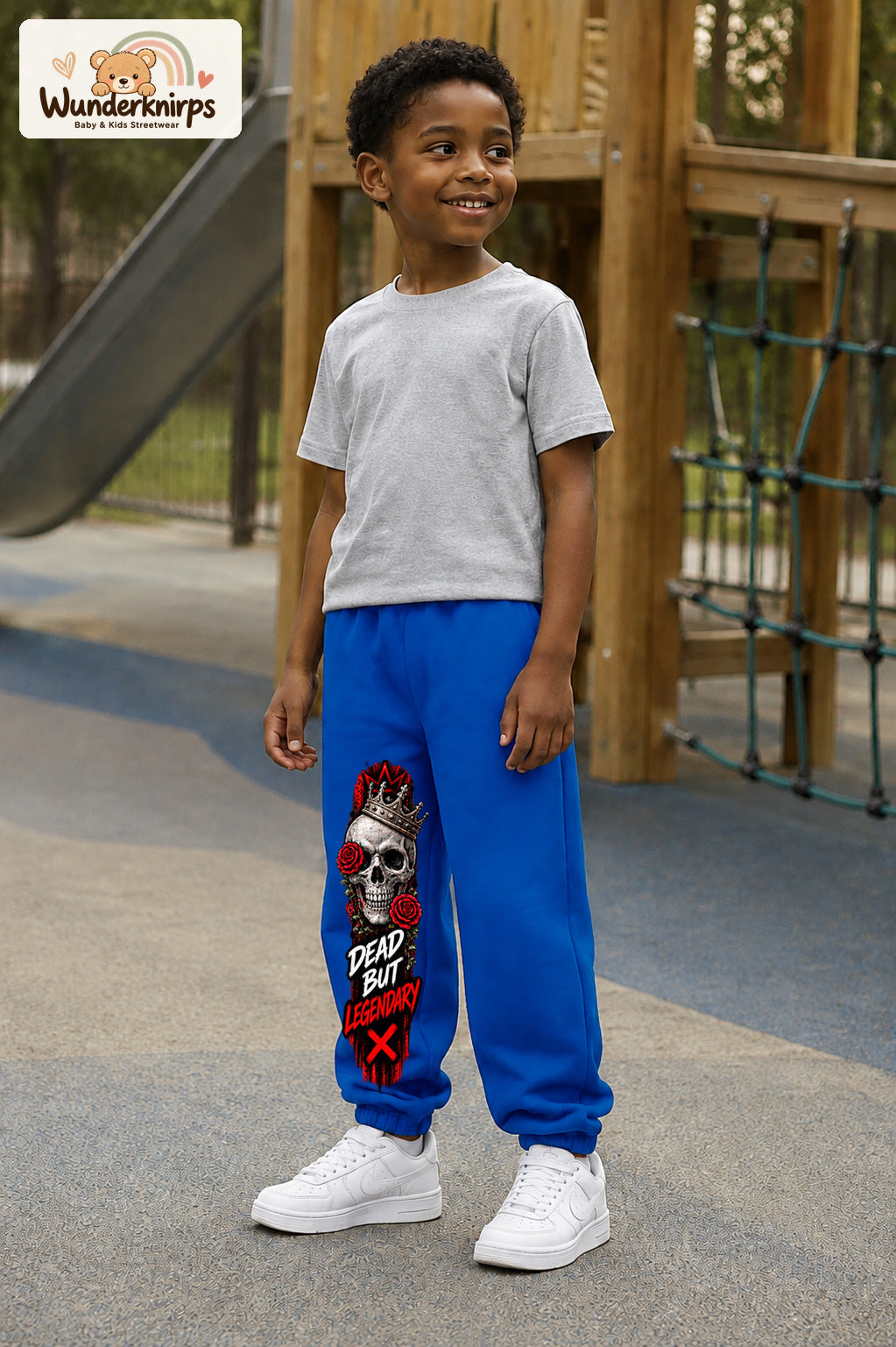 Junge trägt blaue Jogginghose mit Dead but Legendary-Print, leicht seitlich auf dem Spielplatz.