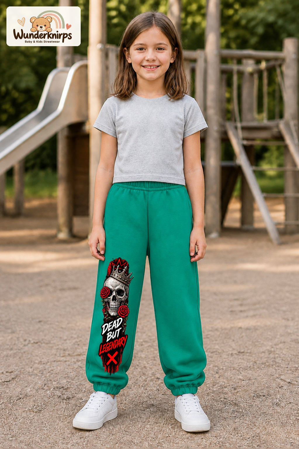 Mädchen trägt grüne Jogginghose mit Dead but Legendary-Print, von vorn auf dem Spielplatz.